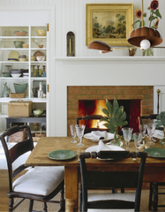 Warm dining room featuring the Fudge Brown & Brass Coquille chandelier with scalloped shell-inspired shades, suspended above a rustic wood dining table set with green plates, crystal glassware, and white linens. The cozy space includes a brick fireplace, vintage ladder-back chairs, open shelving with pottery, and framed artwork, blending artisanal statement lighting with traditional farmhouse charm.
