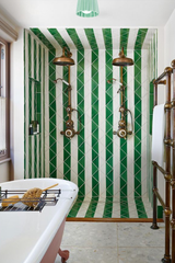 Luxurious green and white striped tile bathroom featuring vintage brass double shower fixtures and a clawfoot tub with pink exterior. Overhead, a handmade jade green ceramic flush mount ceiling light in an Art Deco-inspired scalloped design adds sculptural elegance and a pop of pastel contrast. The bold geometric tile pattern creates a visually striking space, while the artisanal ceramic light fixture softens the room with handcrafted detail. Perfect inspiration for maximalist, vintage-inspired bathroom dec
