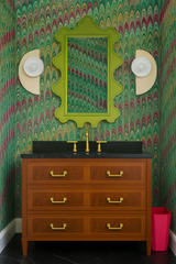 Bold, eclectic green bathroom with vibrant marbled wallpaper in pink, green, and yellow tones, featuring a chartreuse scalloped mirror and a pair of color-blocked Kin Sconces in cream and white, flanking a warm wood vanity with brass hardware and black stone countertop.