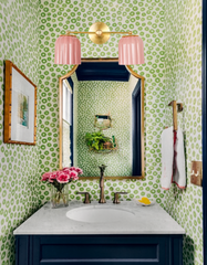 Vibrant powder room featuring the Double Marcella Sconce with ribbed pastel pink ceramic shades and a warm brass backplate, mounted above a marble vanity with navy cabinetry and brass faucet. The bathroom is styled with bold green spotted wallpaper, a gold-trimmed mirror, and fresh pink flowers, showcasing how handmade ceramic wall lighting by Sazerac Stitches adds playful Art Deco-inspired elegance and a pop of color to eclectic, modern interiors.