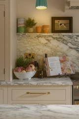 The Marcella sconce in glossy jade green ceramic with a scalloped edge glows warmly above a kitchen counter styled with dramatic marble backsplash, cream cabinetry with brass hardware, a cookbook on display, a bowl of fresh root vegetables, and a small herb plant—creating an inviting, lived-in feel perfect for eclectic kitchens or English countryside–inspired interiors.