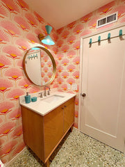 Poolside blue Clancy sconce with polished brass hardware brightens a retro-inspired bathroom with pink and orange fan-patterned wallpaper, terrazzo flooring, and a warm oak vanity. This midcentury wall light by Sazerac Stitches brings a cheerful, colorful accent that makes small bathrooms or powder rooms feel artful and custom.