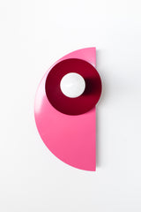 Kin Sconce in a bold palette of doll pink, glossy black cherry, and matte terra cotta, shown mounted on a white wall. The sculptural silhouette and layered color give this geometric fixture a modern, Bauhaus-inspired look.