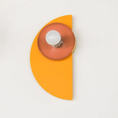 A graphic, Bauhaus-inspired wall sconce in a sunny mustard yellow half-circle, paired with a warm peach disk and olive green socket. This playful fixture adds a splash of Memphis-modern charm to maximalist, retro, or artsy interiors.