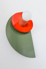 Maximalist color-blocked Kin Sconce with a powder-coated olive green semicircle backplate, glossy flame red dish, and muted peach socket, topped with a white globe bulb and mounted on a brass backplate for an eclectic modern look.