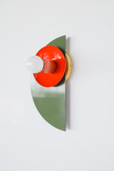 Maximalist color-blocked Kin Sconce with a powder-coated olive green semicircle backplate, glossy flame red dish, and muted peach socket, topped with a white globe bulb and mounted on a brass backplate for an eclectic modern look.