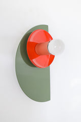 Maximalist color-blocked Kin Sconce with a powder-coated olive green semicircle backplate, glossy flame red dish, and muted peach socket, topped with a white globe bulb and mounted on a brass backplate for an eclectic modern look.