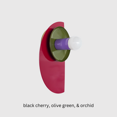 A bold Kin Sconce in a striking trio of black cherry, olive green, and orchid purple. The glossy black cherry half-moon backplate grounds the fixture, while a mossy olive dish and lavender-toned socket create a high-contrast, playful color story. Perfect for eclectic spaces, maximalist interiors, or color-forward homes that lean into Bauhaus or Memphis design cues.