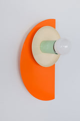 Bright and modern geometric wall sconce in a color-blocked palette of orange, cream, and mint green, shown against a white wall. The fixture includes a layered combination of semicircle and circular steel forms in a sculptural, midcentury-inspired design.