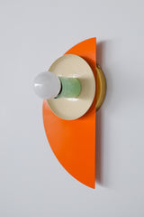 Wall-mounted Kin Sconce featuring a bold orange semicircle backplate, glossy cream round reflector dish, and pastel mint green cylindrical socket cup, all crafted from powder-coated steel and topped with a round white globe bulb.