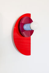 Angled wall-mounted view of the Kin Sconce in a bright red, plum, and blue color-block palette, showing off its layered geometric design.