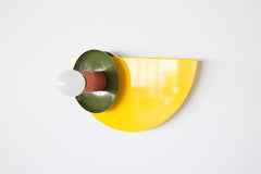 Colorful Kin wall sconce featuring a glossy yellow powder-coated backplate, olive green dish, and terra cotta socket, photographed against a white wall—ideal for maximalist, eclectic, or midcentury modern interiors seeking a pop of color and playful geometric wall lighting.