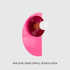 Colorful Kin Sconce featuring bold doll pink, deep black cherry, and warm terracotta tones in a geometric, half-moon design—ideal for eclectic interiors, maximalist bedrooms, or color-drenched powder rooms. The contemporary wall light includes a white globe bulb, layered powder-coated steel plates, and a brass backplate, blending postmodern style with playful, monochromatic color blocking.