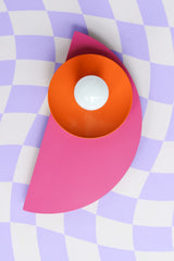An eye-catching Kin Sconce in a vibrant trio of doll pink, orange peel, and hydrangea blue, set against a whimsical lavender and white warped checkerboard background. The glossy finishes and saturated hues lend a retro-modern energy that works beautifully in Memphis, postmodern, or colorful maximalist spaces. This wall light functions as both a statement piece and functional lighting for bedrooms, bathrooms, or studio nooks.