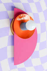 Memphis meets modern in this dynamic Kin Sconce, shown in a saturated palette of doll pink, orange peel, and hydrangea blue. The half-circle backplate and nested disc form a geometric composition that nods to Bauhaus principles with a pop-art twist. Set against a distorted lilac checkerboard surface, this piece is perfect for playful interiors, gallery walls, or anywhere bold design is welcome.