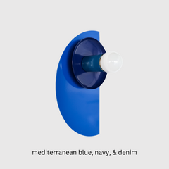 A sculptural Kin Sconce in a tone-on-tone palette of Mediterranean blue, navy, and denim, mounted on a clean white wall. The deep navy shade and denim socket sit against a glossy half-moon backplate in vibrant Mediterranean blue, creating a layered, Bauhaus-inspired silhouette. The rich monochromatic color scheme gives this geometric fixture a bold, modern presence that works beautifully in eclectic interiors or maximalist spaces.