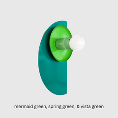 A sculptural Kin Sconce in a tone-on-tone palette of Mermaid Green, Spring Green, & Vista Green, mounted on a clean white wall. The spring green shade and pastel green socket cup sit against a glossy half-moon backplate in vibrant Mermaid Green, creating a layered, Bauhaus-inspired silhouette. The rich monochromatic color scheme gives this geometric fixture a bold, modern presence that works beautifully in eclectic interiors or maximalist spaces.