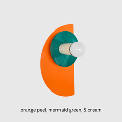 The Kin Sconce in a juicy orange, high-gloss mermaid green, and soft cream socket creates a playful, color-blocked statement. The bright semicircle backplate delivers punchy contrast against the deep teal-green dish and neutral socket. This fixture brings geometric charm to retro-inspired, maximalist, or Bauhaus-influenced interiors—and works beautifully as a bold bathroom sconce.