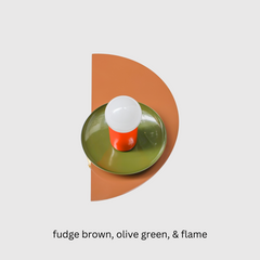 A geometric Kin Sconce in rich fudge brown, glossy olive green, and flame red, shown against a white wall. The half-circle backplate in warm brown supports a green shade and bold red socket, forming a layered Bauhaus-inspired silhouette. This sculptural light blends earthy and modern tones, making it ideal for contemporary kitchens, reading nooks, or color-forward living spaces.