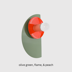 Maximalist color-blocked Kin Sconce with a powder-coated olive green semicircle backplate, glossy flame red dish, and muted peach socket, topped with a white globe bulb and mounted on a brass backplate for an eclectic modern look.
