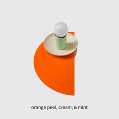 Bright and modern geometric wall sconce in a color-blocked palette of orange, cream, and mint green, shown against a white wall. The fixture includes a layered combination of semicircle and circular steel forms in a sculptural, midcentury-inspired design.