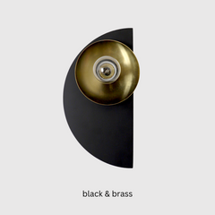 The Kin Sconce in matte black and polished brass features a bold semicircle backplate paired with a gleaming brass shade and socket. With its crisp geometric form and sculptural silhouette, this wall light is a nod to Bauhaus and Memphis design principles—clean lines, striking shapes, and elevated simplicity. The contrast of deep black and warm metallic makes it a standout accent in both minimal and maximalist interiors. Ideal for powder rooms, hallway vignettes, or flanking a bed in a modern, transitional,