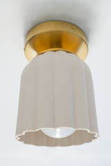 Cream-colored unglazed porcelain flushmount ceiling light illuminated from within, glowing warmly through its translucent ceramic shade. The handmade fixture features sculptural fluted ridges and a scalloped bottom edge, paired with a sleek brass canopy. Its Art Deco-inspired silhouette and soft ambient glow make it ideal for hallways, powder rooms, and layered lighting in colorful, design-forward homes