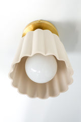 Cream-colored unglazed porcelain flushmount ceiling light illuminated from within, glowing warmly through its translucent ceramic shade. The handmade fixture features sculptural fluted ridges and a scalloped bottom edge, paired with a sleek brass canopy. Its Art Deco-inspired silhouette and soft ambient glow make it ideal for hallways, powder rooms, and layered lighting in colorful, design-forward homes