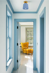 Bright hallway with white walls and powder blue trim, featuring a handmade ceramic flush mount ceiling light in a matte soft blue finish with brass hardware. The scalloped Art Deco-inspired shade adds a pop of subtle color and sculptural charm. Through the open doorway, a vibrant mustard yellow tufted armchair brings a warm contrast to the cool tones, creating a cheerful and inviting space filled with natural light and bold color accents. Perfect lighting inspiration for modern cottage or color-drenched int