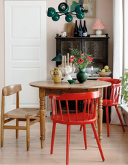Eclectic dining room with the Loa Carousel chandelier in mermaid green and black, featuring sculptural globe shades above a round wooden dining table. The space includes bold red dining chairs, vintage wood cabinet with patterned lamp, fresh flowers, and playful ceramics, blending colorful statement lighting with cozy modern cottage style.