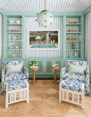 Coastal-inspired sitting room featuring the Mint & Brass Gumball pendant light, a round globe fixture with sculptural mint accents, hanging from a brass stem. The space includes blue and green patterned armchairs with matching ottomans, striped wallpaper, mint built-in shelving, and woven side tables, blending playful statement lighting with breezy, beach house interior design.