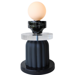 Sculptural table lamp glowing warmly with a frosted glass globe on top of a reclaimed black marble element, thick clear acrylic disc, and fluted matte black ceramic base; unique ambient lighting fixture handmade in New Orleans, perfect for a modern eclectic or artful interior