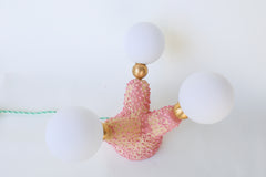 Angled photo of a unique handmade ceramic table lamp resembling an abstract coral or sea creature, built from stoneware and finished in layered pink glazes; features three bulb arms with brass hardware and frosted G9 globes, perfect for ambient mood lighting in a quirky living room, bedroom, or artistic shelf display.