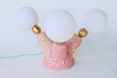 A one-of-a-kind handmade pink ceramic table lamp with a creature-inspired silhouette featuring three organically shaped arms glazed in bubblegum pink with pastel pink undertones, finished with raw brass ball joints, frosted white G9 screw-on glass globes, and a mint green plug-in cord with rocker switch—ideal for offbeat, whimsical, and maximalist home decor.