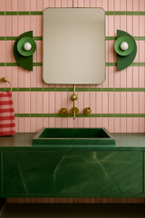 Bold, color-forward bathroom with a striking green marble vanity and vertically stacked blush pink tiles accented with olive green grout lines. A pair of Kin Sconces in coordinating green hues flank a rectangular mirror above a brass wall-mounted faucet. The playful color palette and geometric lighting add a whimsical yet polished touch to this modern powder room or kids’ bathroom design.