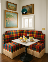 Colorful breakfast nook featuring the Large Loa pendant light in mermaid green and brass, suspended above a marble café table with a black cast-iron base. The corner banquette is upholstered in bold plaid fabric with fringe trim, paired with framed vintage paintings, croissants, coffee, and fresh fruit, blending playful retro lighting with eclectic cottage-inspired dining design.