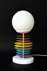 The Vertebrale lamp by Sazerac Stitches combines color, form, and functionality. Powder-coated steel discs in bold hues, a glowing frosted globe, and a sleek marble base create a striking modern design ideal for any interior.