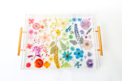 Handmade acrylic tray featuring real dried and pressed flowers arranged in a rainbow gradient layout, with modern gold handles on either side. The flowers are suspended in clear resin, creating a vibrant, colorful design that is perfect for spring decor. Ideal for use as a vanity tray, coffee table tray, or bar accessory in bright, eclectic, or cottage-inspired interiors.