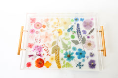 Handmade acrylic tray featuring real dried and pressed flowers arranged in a rainbow gradient layout, with modern gold handles on either side. The flowers are suspended in clear resin, creating a vibrant, colorful design that is perfect for spring decor. Ideal for use as a vanity tray, coffee table tray, or bar accessory in bright, eclectic, or cottage-inspired interiors.