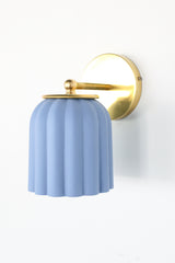 Unglazed soft blue ceramic wall sconce with solid brass hardware, featuring a scalloped silhouette that brings a charming touch to bathroom and powder room lighting; ideal for traditional, coastal, Nantucket-inspired, Southern, or transitional interiors where soft color and timeless form add a refined yet playful focal point.