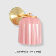 Close-up of the Marcella ceramic wall sconce in glazed pastel pink with a fluted shade and satin brass backplate, shown against a neutral background. This whimsical yet refined light fixture blends playful retro charm with elevated modern design, perfect for traditional, coastal, or cottagecore interiors seeking a soft pop of color and timeless silhouette.