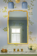 Elegant powder room styled as a jewel-box retreat, featuring the Double Marcella Sconce in glossy sky blue ribbed ceramic shades with a warm brass backplate above a marble vanity. The bathroom showcases a traditional Southern design aesthetic with hand-painted floral wallpaper in soft blue and green, a gilded mirror, brass faucet fixtures, and fresh white hydrangeas. Perfect for classic, traditional homes or luxury Southern interiors, this sophisticated wall sconce by Sazerac Stitches adds Art Deco charm an