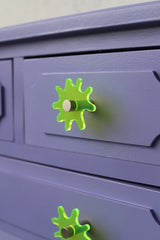 Splat drawer pull in translucent neon green acrylic with a brass detail installed on a colorful lavender dresser drawer, demonstrating its easy installation and ability to add a unique touch.