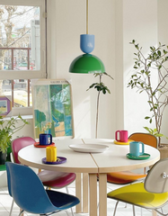Bright, modern dining room featuring the Taeuber pendant light in blue and green, suspended above a round white dining table surrounded by mismatched colorful chairs. The eclectic space includes potted plants, framed artwork, and bold multicolor mugs, blending playful retro-inspired lighting with vibrant contemporary interior design.