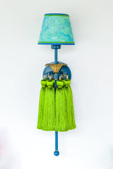 Straight-on shot of the Tropical Tasseled Wall Sconce featuring lagoon blue metalwork, antique brass detailing, chartreuse tassels, and an aqua marbled lampshade. This playful, statement-making designer light adds color-drenched, tropical-inspired flair to hallways, bedrooms, or eclectic living spaces.