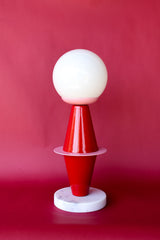 The Totem Lamp by Sazerac Stitches combines bold geometric shapes with Bauhaus-inspired design. Crafted from powder-coated steel in vibrant red and pink hues, it features a frosted globe and sleek marble base.