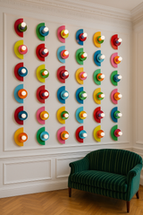 A wall of colorful Kin Sconces arranged in a 7x5 grid in a classic interior with white panel molding. The vibrant geometric fixtures in bold color combinations pop against the neutral backdrop, with a green velvet loveseat positioned below.