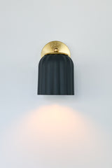 Marcella wall sconce with unglazed matte black ceramic shade and brushed brass backplate, shown on a neutral background. The scalloped silhouette and soft finish evoke timeless elegance with a modern edge, perfect for moody interiors, modern traditional decor, or eclectic spaces seeking contrast and sophistication.