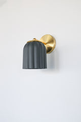 Marcella wall sconce with unglazed matte black ceramic shade and brushed brass backplate, shown on a neutral background. The scalloped silhouette and soft finish evoke timeless elegance with a modern edge, perfect for moody interiors, modern traditional decor, or eclectic spaces seeking contrast and sophistication.