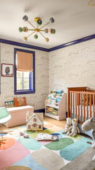 Modern kids’ nursery with Orion Chandelier in cream, grey, and vista by Sazerac Stitches. Room features colorful geometric rug, midcentury rocking chair, wood crib, toy animals, and book storage under navy blue window trim. Stylish midcentury modern nursery lighting.